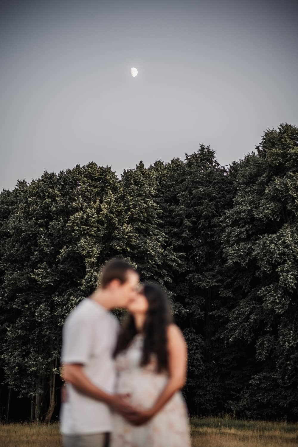 Schwangere Mama mit Partner unscharf im Vordergrund. Im Hintergrund Wald und Mond beim Babybauch-Shooting.