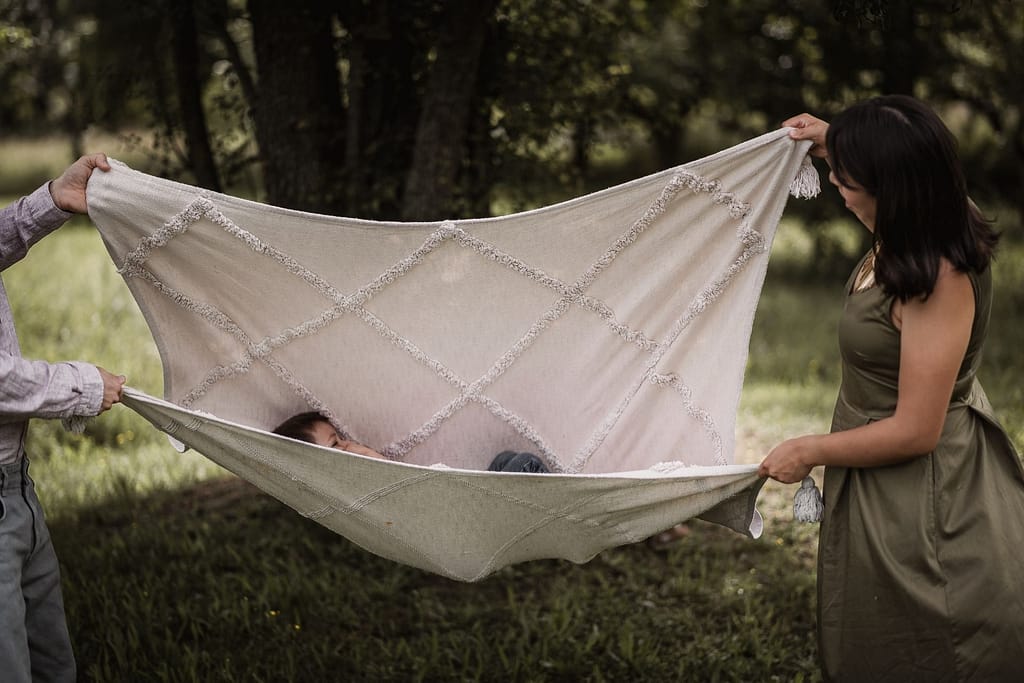 Mama und Papa halten eine cremefarbene Picknickdecke als Schaukel. In der Decke liegt ihr 3-jähriger Sohn, der sich beim Familienshooting schaukelnlässt.