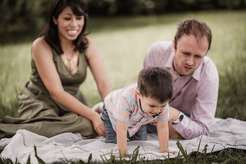 Beim Familienshooting lachen Mama und Papa mit ihrem 3-jährigen Sohn. Der Junge erkundet etwas im Gras.