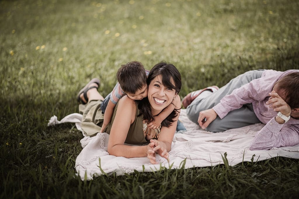 Beim Familienshooting lachen Mama und Papa mit ihrem 3-jährigen Sohn. Der Junge liegt auf dem Rücken seiner Mama, die in die Kamera lächelt.