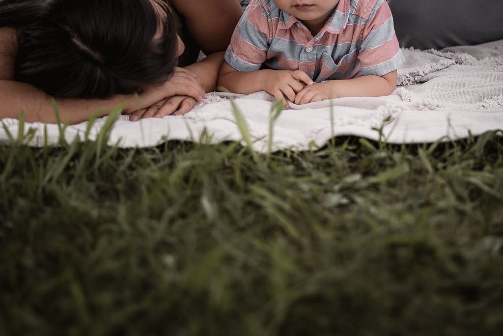 Detailaufnahme von Mama und ihrem 3-jährigen Sohn, der beim Familienshooting neben ihr auch einer hellen Picknickdecke liegt. Man sieht nur ihre Hände.