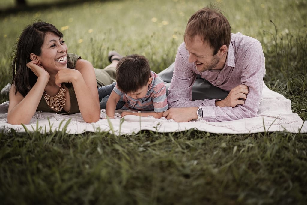 Beim Familienshooting lachen Mama und Papa mit ihrem 3-jährigen Sohn. Der Junge erkundet etwas im Gras.