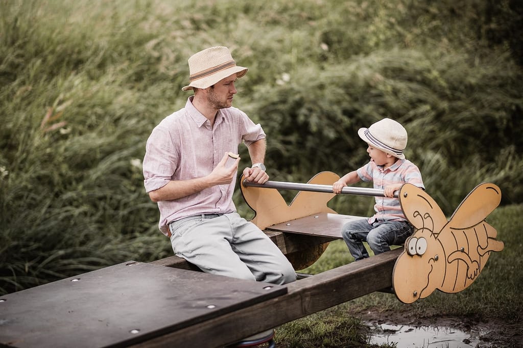 Papa und Sohn beim Familienshooting. Sie sitzen auf einer Wippe. Beide haben Stohhüte auf und schauen sich an.