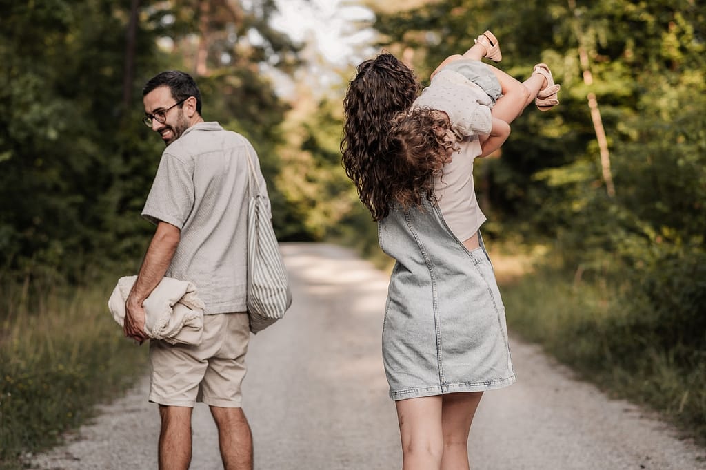 Mama und Papa laufen auf Waldweg. Ihr KLeinkind hängt lustig über der Schulter. Papa schaut zurück.