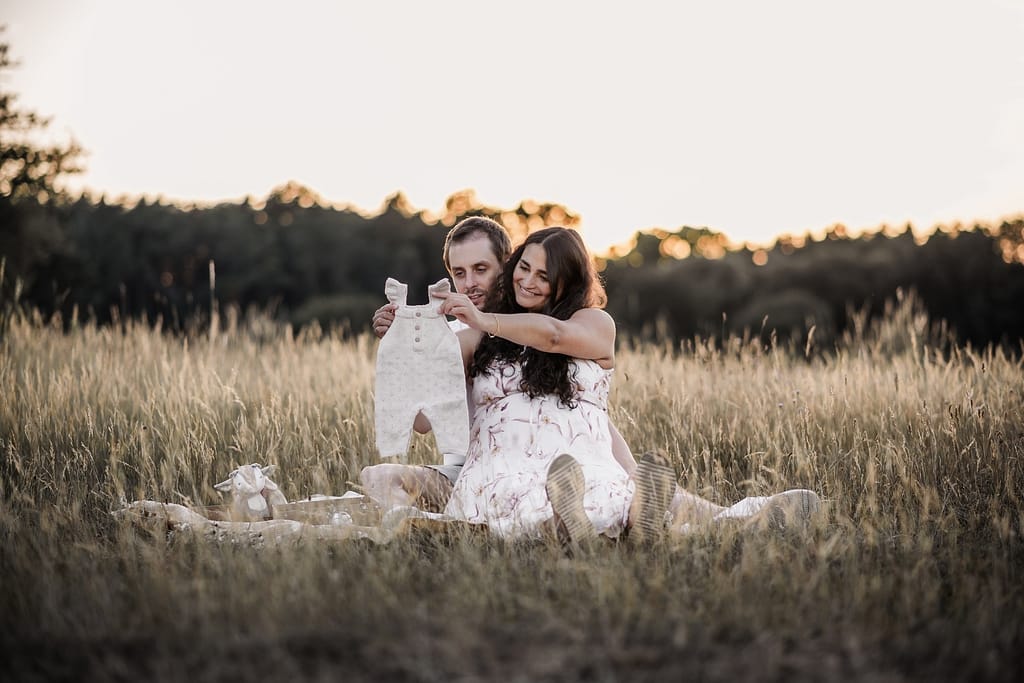 Werdende Eltern beim Babybauch-Shooting sitzen in der Wiese und genießen Zeit zusammen und betrachten die Baby-Kleidung.
