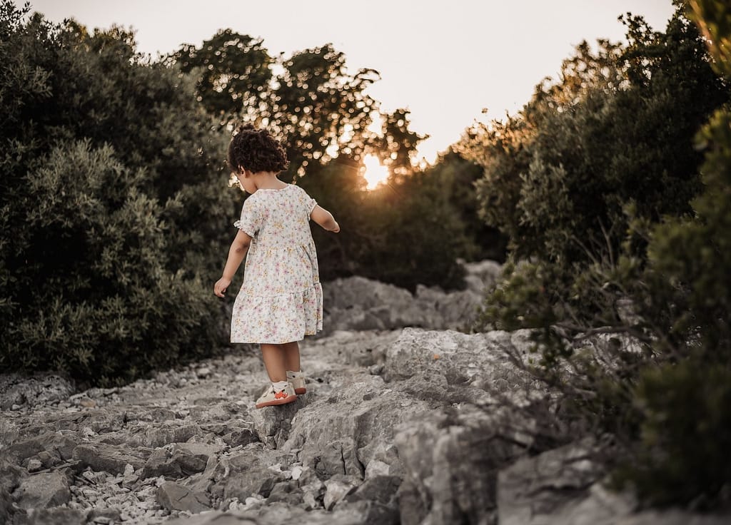 Kleinkind in einem hellen Kleid in der Natur bei Sonnenuntergang zwischen Wald und Steinen.