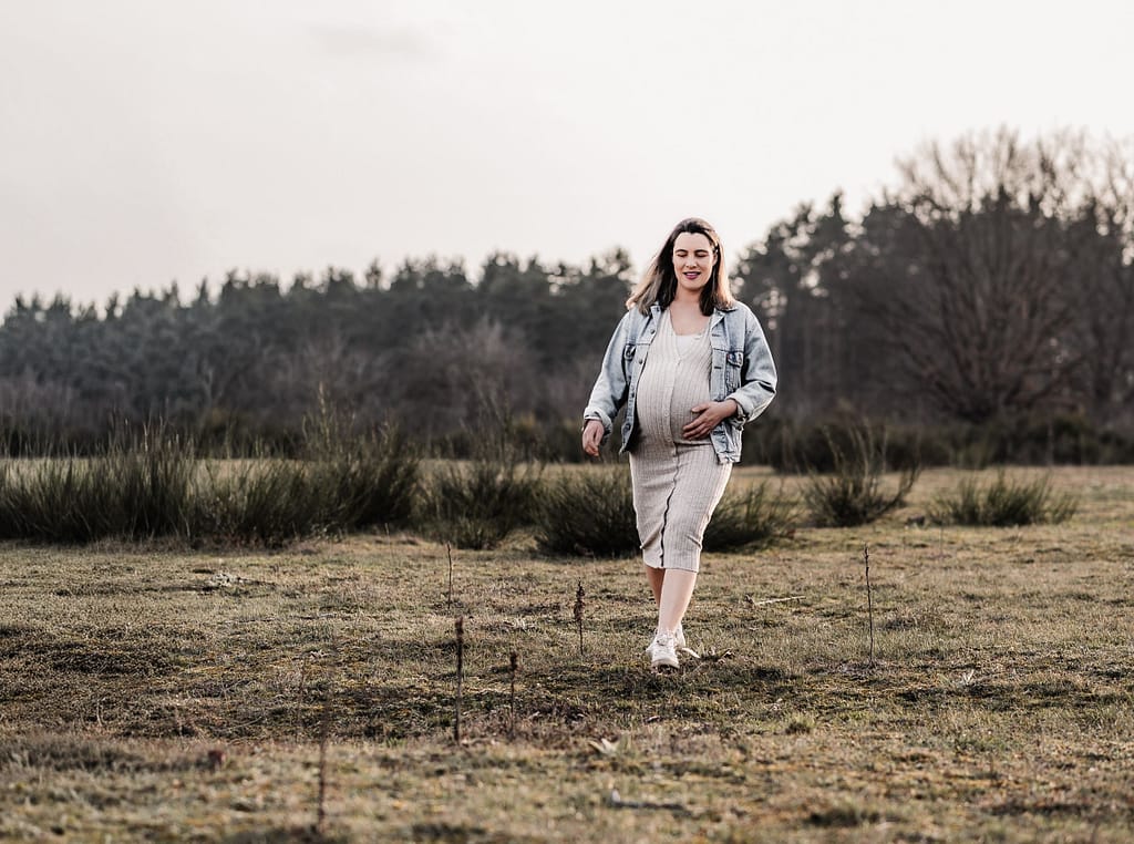Schwangere Frau auf einer Wiese und schaut verträumt in die Ferne, während sie sich über die Wiese geht und sich den Babybauch streichelt.