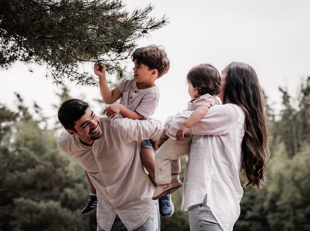Familie mit 2 Kindern unter einer Kiefer. Die Eltern tragen jeweils ein Kind. Der kleine Junge ist auf Papas Rücken und pflückt eine Zapfe vom Baum.