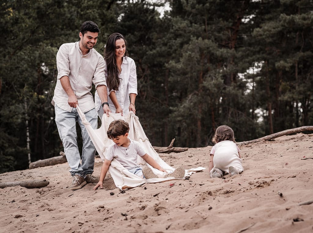 Mama und Papa ziehen ihren Sohn auf einen Picknickdecke durch den Sand. DIe kleine Schwester krabbelt hinterher.