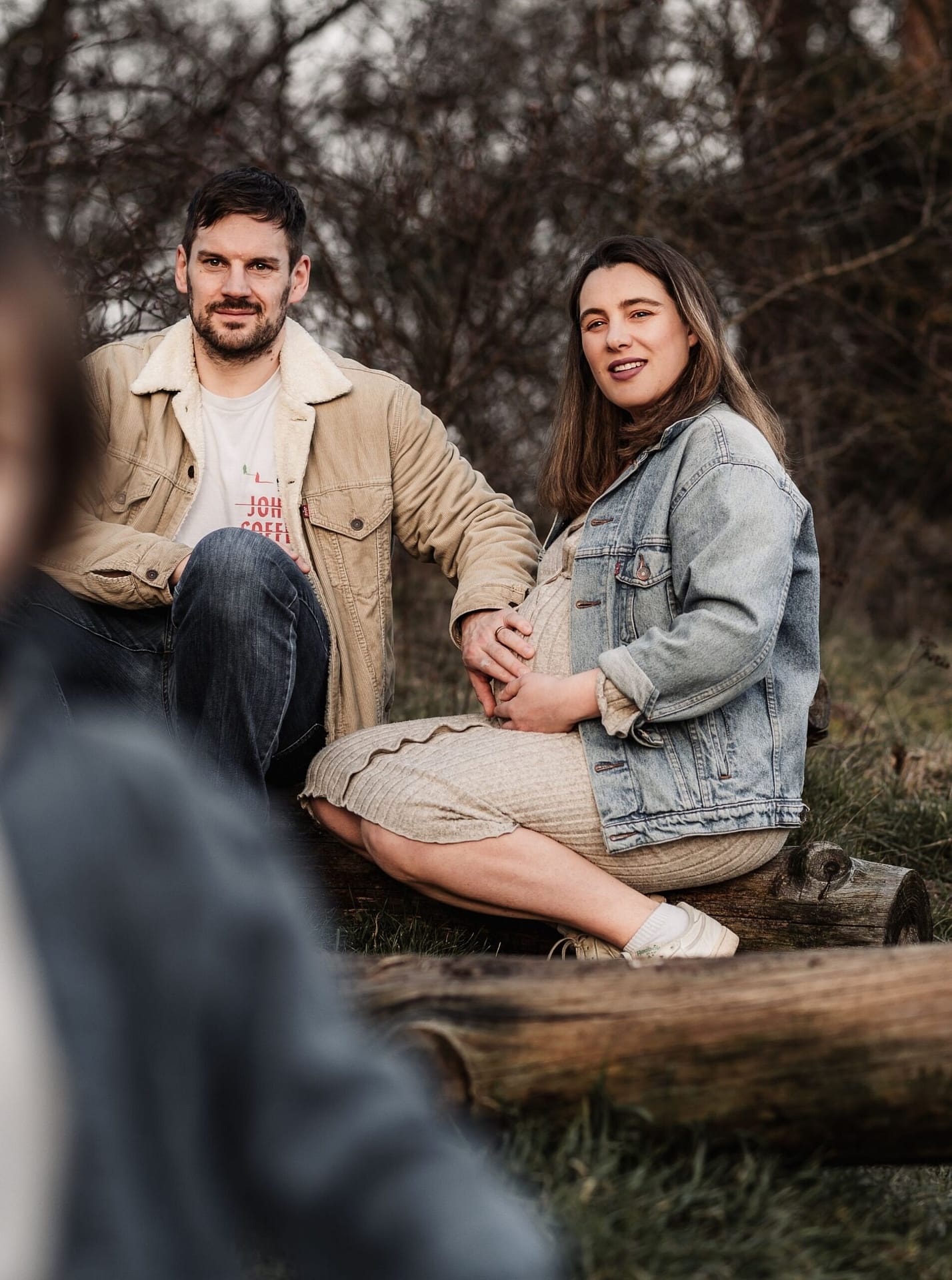 Papa und Mama sitzen auf einem Baumstamm. Die Frau ist schwanger. Papa streichelt zart über den Bauch. Im Vordergrund ist ihr zweijähriger Sohn unscharf zu sehen, der auf die Kamera zurennt.