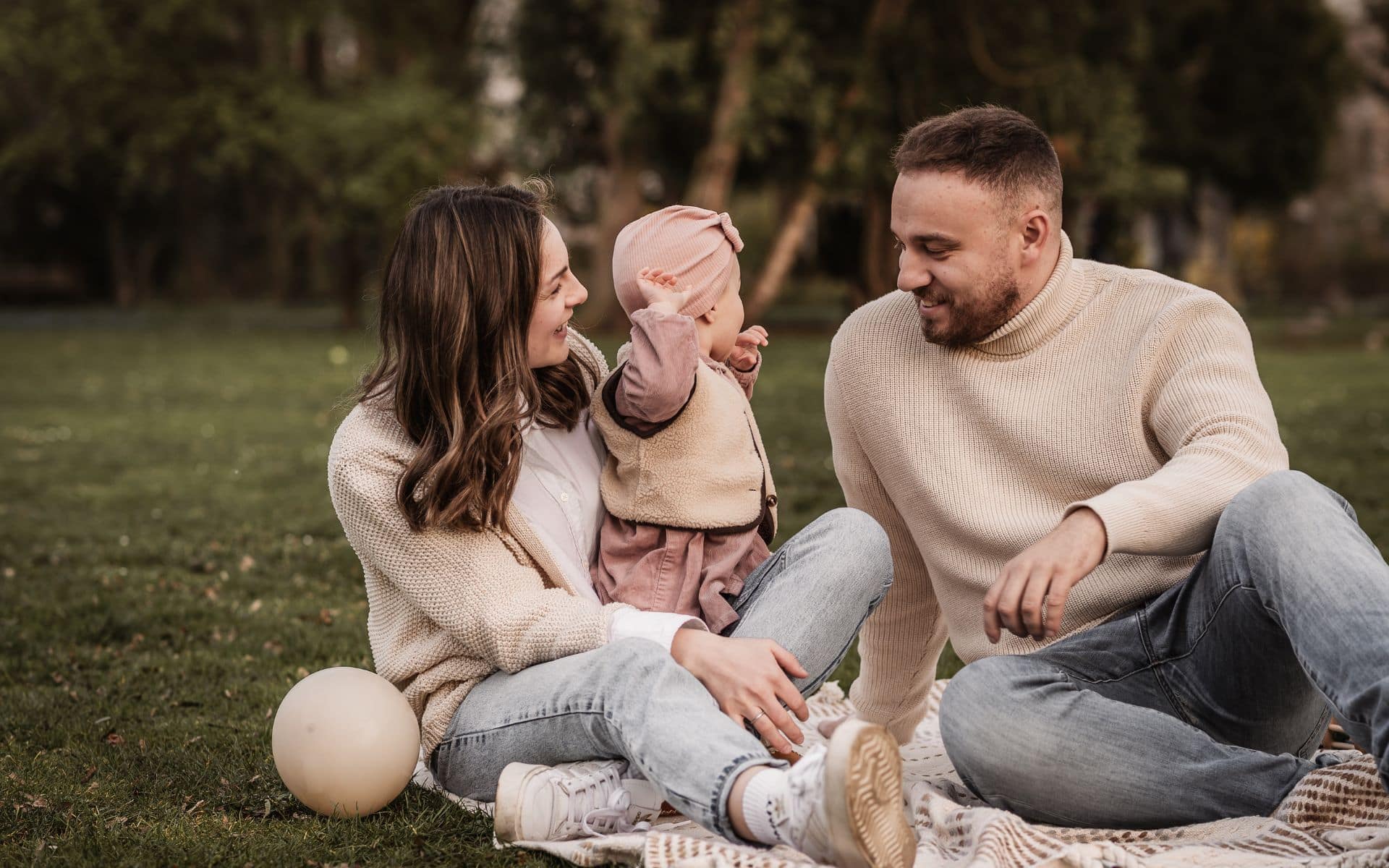 Mama, Papa bei Familienfotos mit Kleinkind im Park auf einer Picknickdecke. Sie lachen miteinander und spielen mit Luftballons.