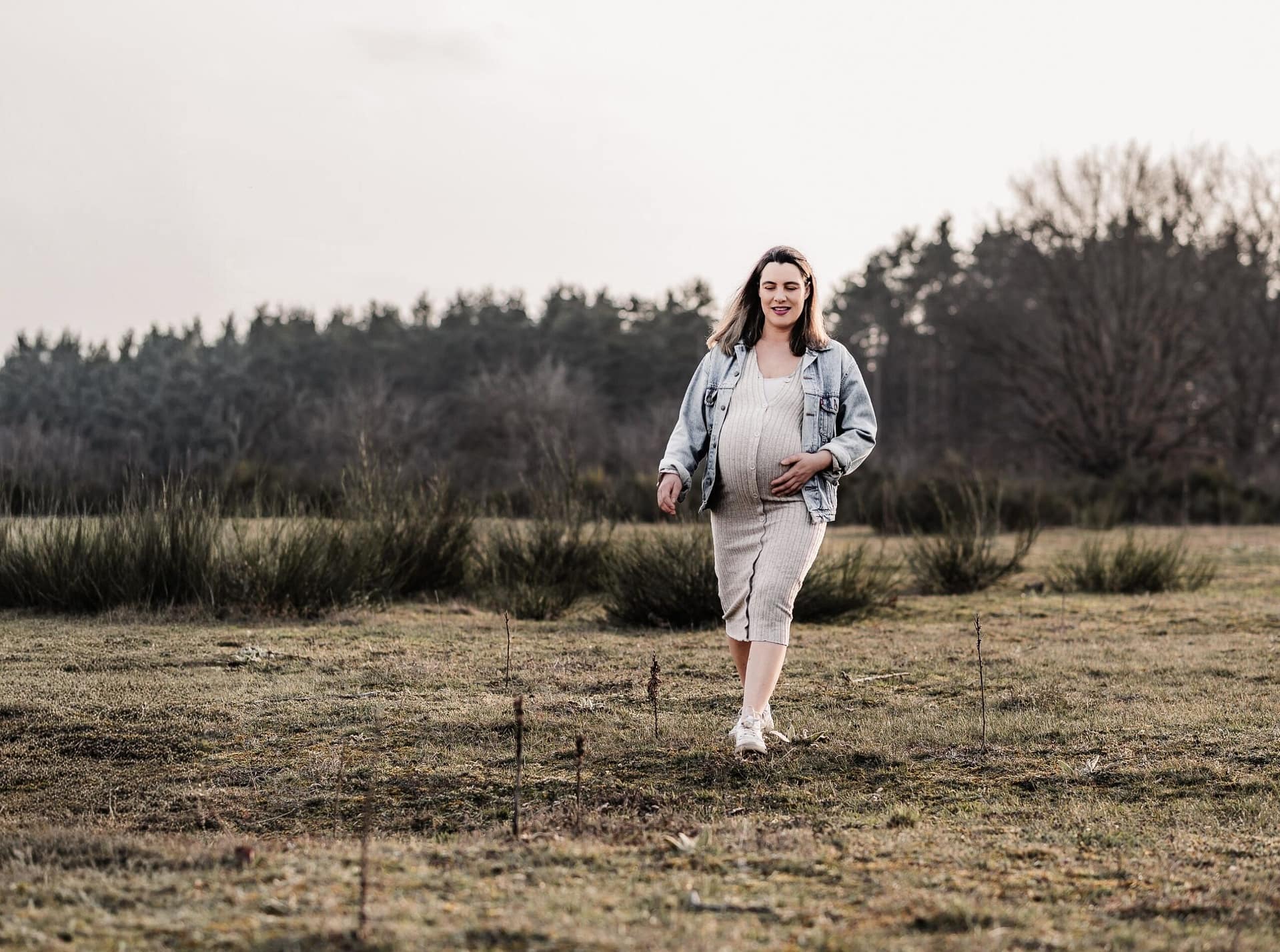 Schwangere Frau auf einer Wiese und schaut verträumt in die Ferne, während sie sich über die Wiese geht und sich den Babybauch streichelt.