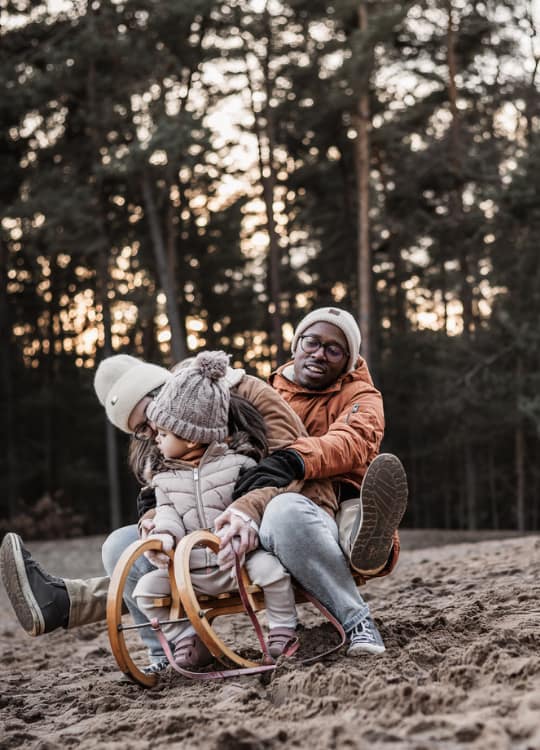 Kleinkind mit Mama und Papa auf Schlitten auf einer Sanddüne bei untergehender Sonne umrahmt von einem Kiefernwald