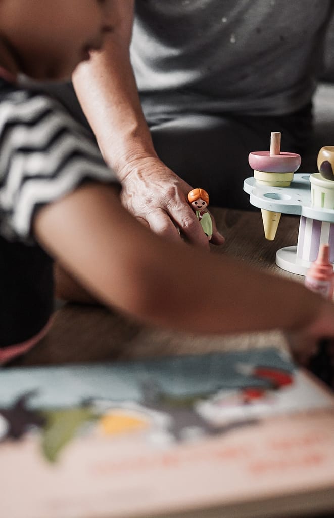 Nahaufnahme von Kleinkind mit Oma beim Spielen. Im Fokus ist eine Spielfigur. Daneben steht ein Holz-Eisständer und im Vordergrund ist ein kleines Kind verschwommen zu sehen.