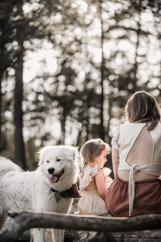 Weißer Hund im Vordergrund, dahinter Mutter und Tochter auf einem Baumstamm im Kiefernwald.