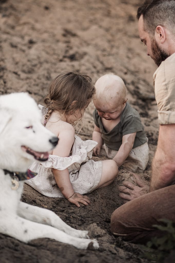 Zwei Kinder spielen mit dem weißen Hund im Sand, der Vater daneben.
