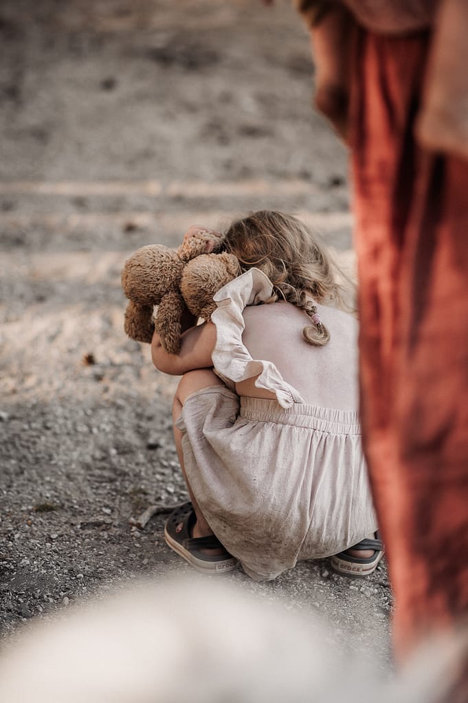 Kleines Mädchen hockt sich mit ihrem Teddybären auf den sandigen Weg.