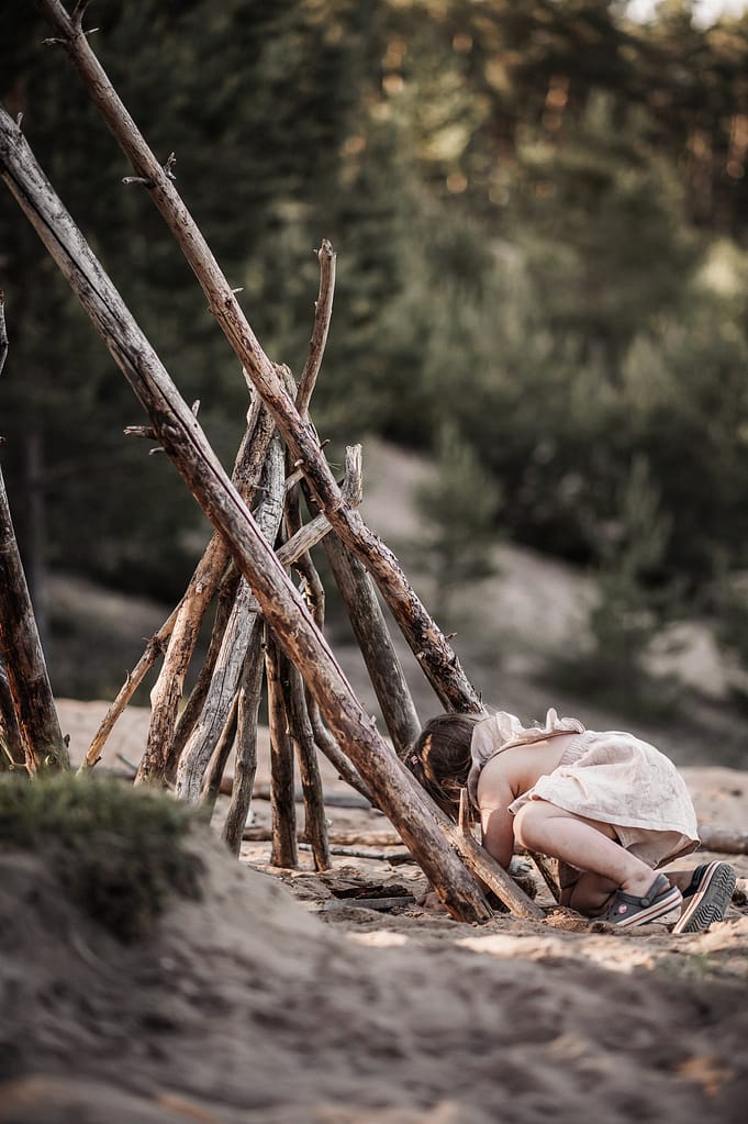Kleines Mädchen sitzt an einem aus Ästen gebauten Tipi im Sand.