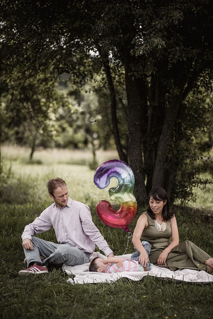 Mama, Papa und kleiner Sohn auf einer Picknickdecke. Der Junge liegt kichernd zwischen seinen Eltern. In der Mitte schwebt ein Folienballon mit der Zahl 3.