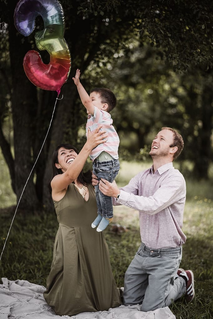 Beim Familienshooting: Mama und Papa heben ihren 3-jährigen Sohn in die Luft. Der Junge strahlt vor Freude während er versucht einen schwebenden Folienballon mit der Zahl 3 zu erreichen.