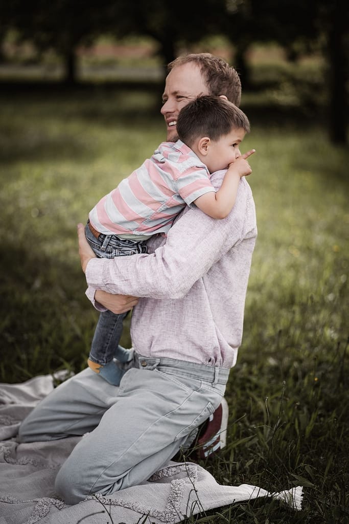 Papa beim Familienshooting auf einer Picknickdecke. Er hält seinen 3-jährigen Sohn auf dem Arm