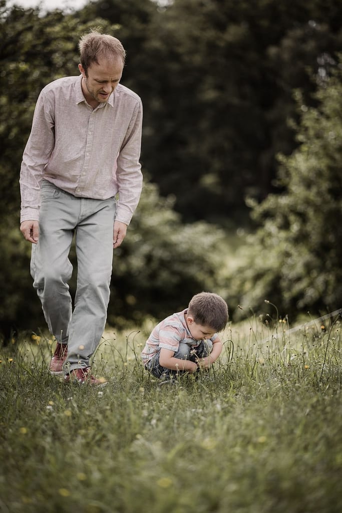 Papa geht beim Familienshooting mit seinem 3-jährigen Sohn über eine Wiese. Der Junge betrachtet etwas im Gras.