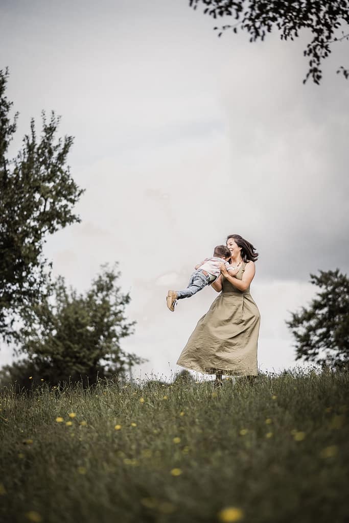 Mama dreht sich mit ihrem 3-jährigen Sohn beim Familienshooting auf einer schönen Wiese im Kreis. Beide lachen.