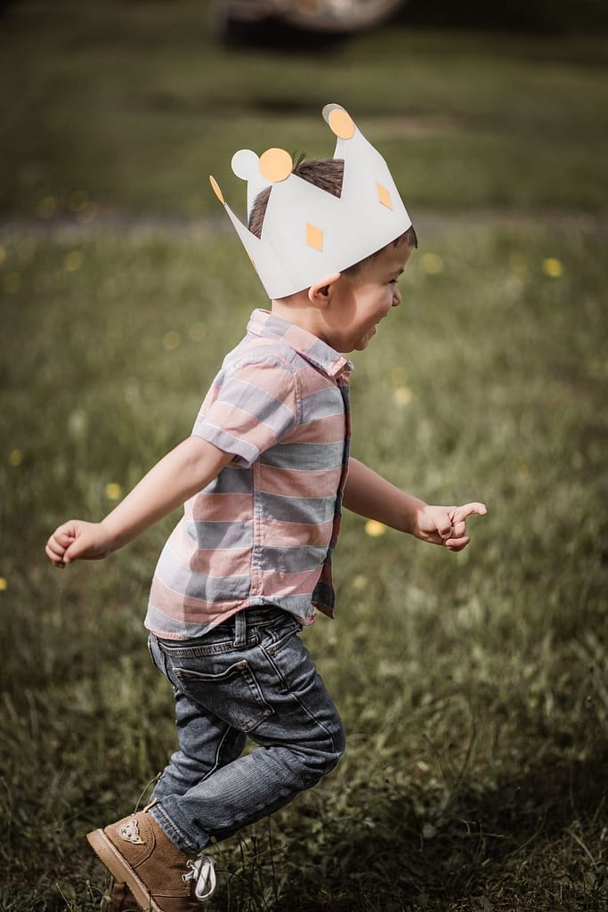 KLeiner Junge rennt beim Familienshooting mit einer Geburtstargskrone über eine schöne Wiese.
