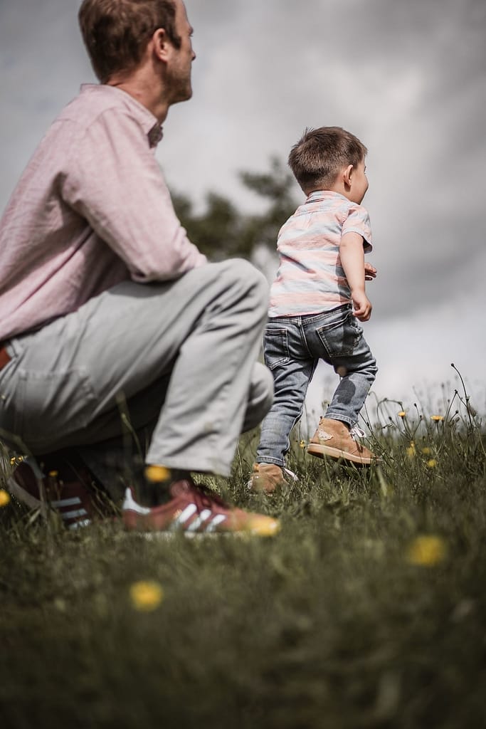 3-jähriger Junge beim Familienshooting rennt von seinem Papa, der auf einer Wiese hockt, los.