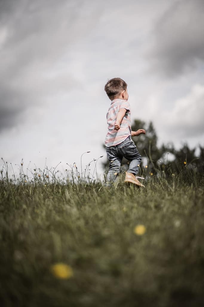 3-jähriger Junge beim Familienshooting rennt über eine grüne Wiese mit Löwenzahl-Blumen weg von der Kamera.