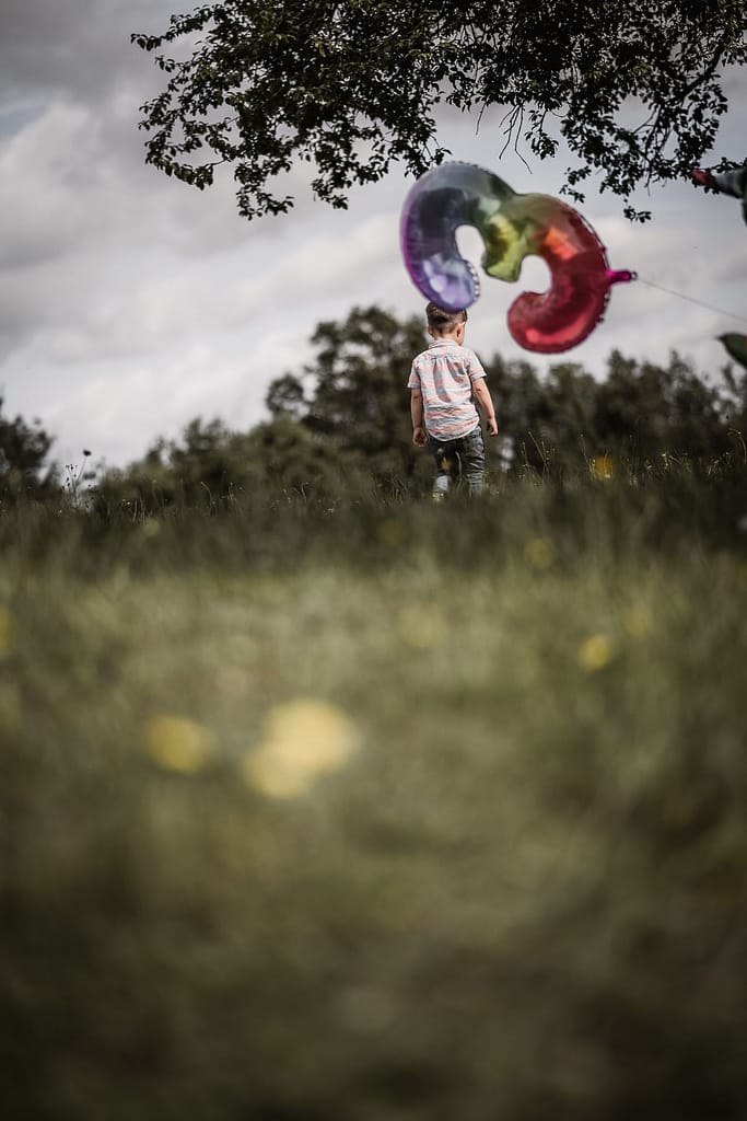 3-jähriger Junge beim Familienshooting rennt über eine grüne Wiese mit Löwenzahl-Blumen weg von der Kamera. Im Vordergrund schwebt ein Folienballon mit der Zahl "3".