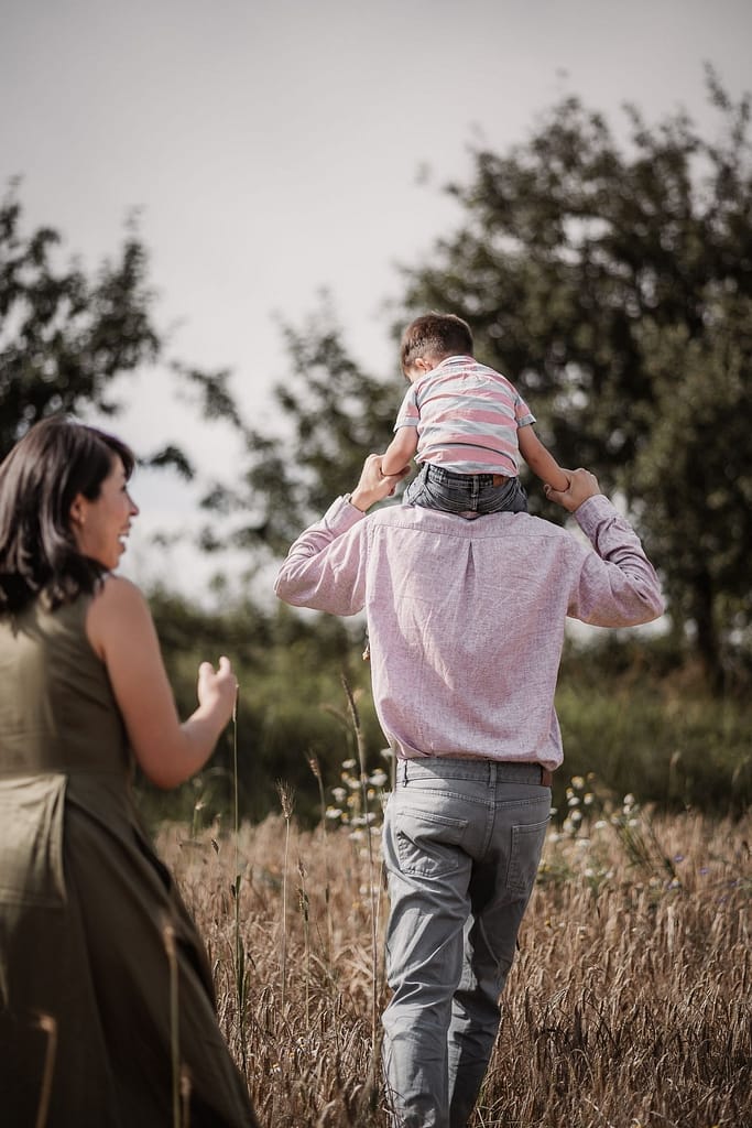 Familienfoto mit Kleinkind. Mama schaut zu, wie Papa ihren Sohn auf den Schultern von durch ein Getreidefeld trägt.