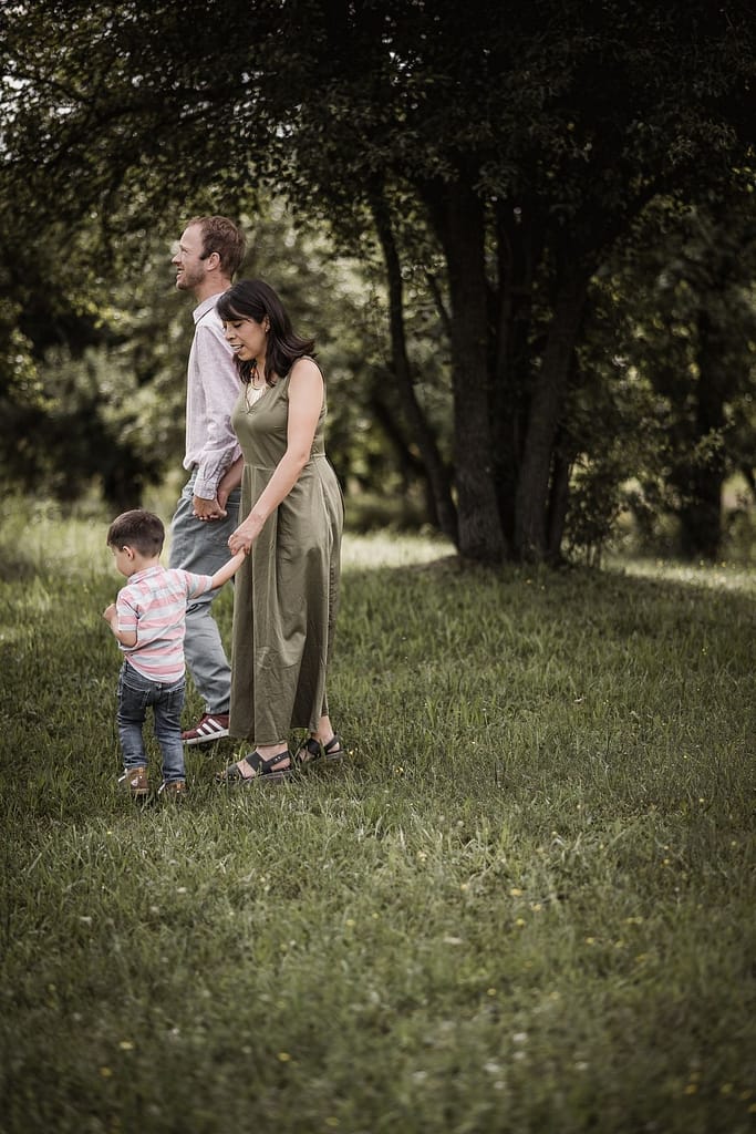 Papa, Mama und Sohn halten sich an den Händen und laufen über eine Wiese inmitten von Apfelbäumen.