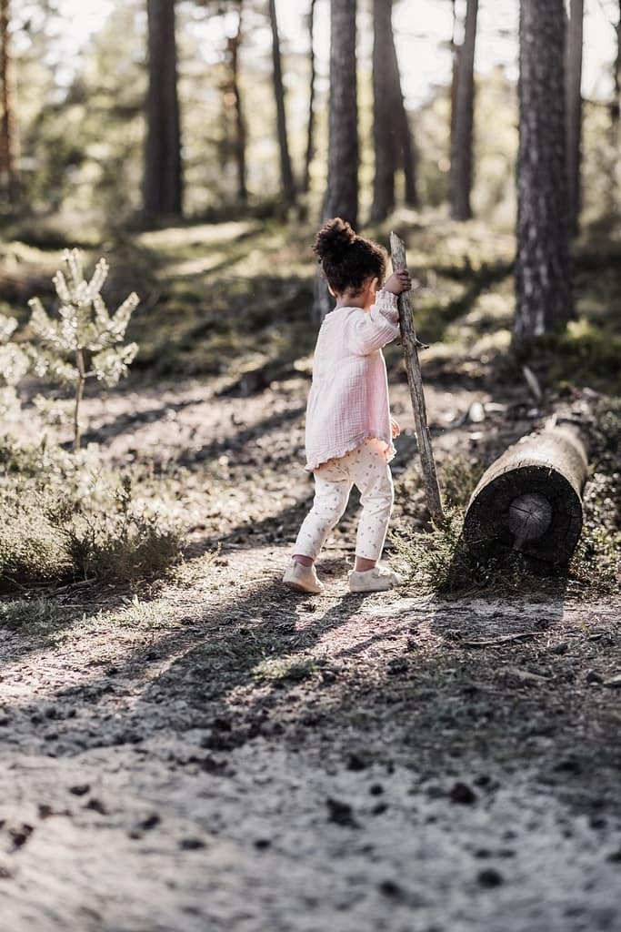 Kleines Kind mit großem Ast als Wanderstock am fränkischen Dünenweg läuft von der Kamera weg Richtung Wald. Zu sehen sind Kiefernwälder im Hintergund im sanften Sonnenlicht und ein sandigen Weg im Vordergrund.