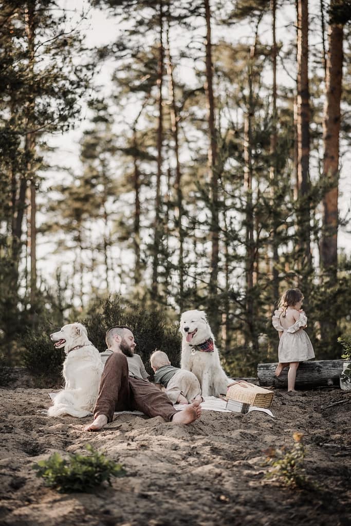 Papa liegt gemütlich auf einer Sanddüne auf einer Picknickdecke. Er iwrkt entspannt. Sein Sohne (1 Jahr) und 3-jährige Tochter bewegen sich um ihn herum. Ein großer weißer Hund sitzt entspannt daneben.