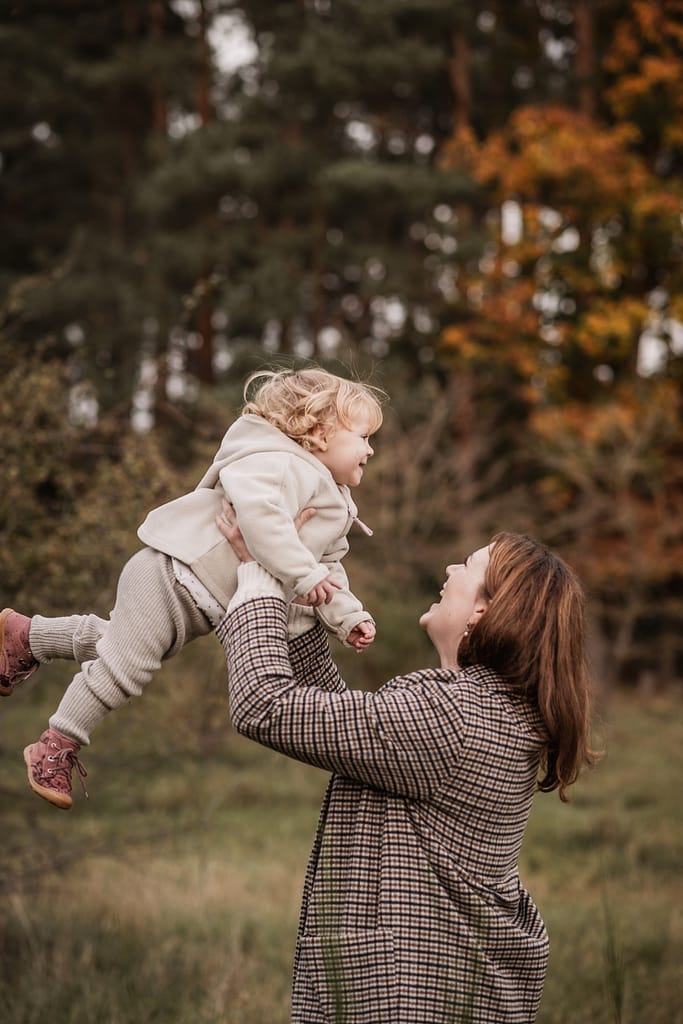 Mama hebt Tochter in die Luft. Sie stehen vor einem Waldstück mit Kiefern und bunten, herbstlichen Bäumen.