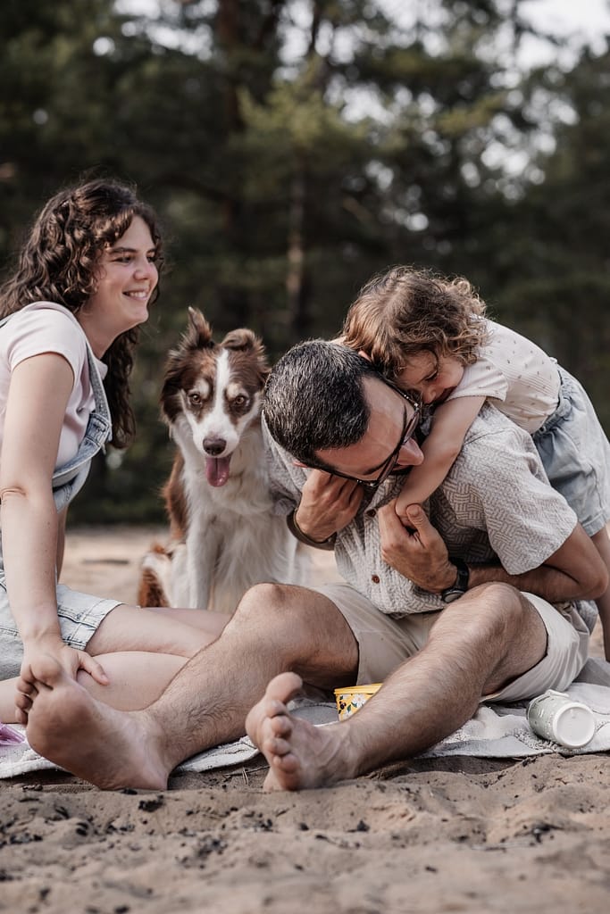Familie mit Hund und KLeinkind am Dünenweg sitz auf einer Picknickdecke beim Familienshooting. Die Tochter drück Papa von hinten und liegt auf seinem Rücken. Alle lachen miteinander.