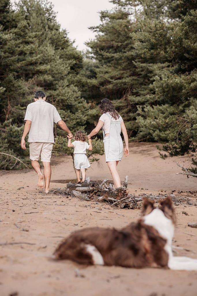 Mama und Papa mit KLeinkind Balancieren auf Baumstamm an Dünenweg. Im Vordergrund liegt ihr Hund und schaut zu.