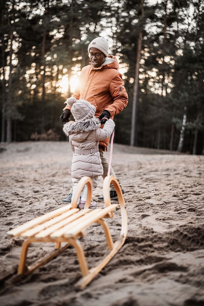 Papa mit Kleinkind und Schlitten auf einer sanddüne mitten Im Wald. Hinter den Bäumen geht die Sonne unter und alles strahlt golden.