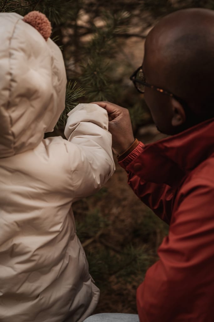 Familienfotos im Winter ohne Schnee mit Papa und KLeinkind. Sie sind von hinter fotografiert und erkunden einen immergrünen Nadelbaum bei Nürnberg.