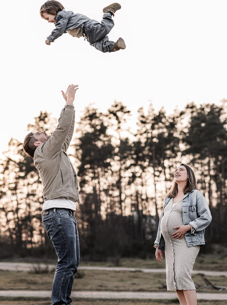Papa wirft zweijährigen Sohn in die Luft während schwangere Mana lächelnd daneben steht. Im Hinterhrund geht die Sonne hinter Kiefernwäldern unter und taucht alles in goldenes Licht beim Schwangerschaftsfotoshooting bei Nürnberg Fürth