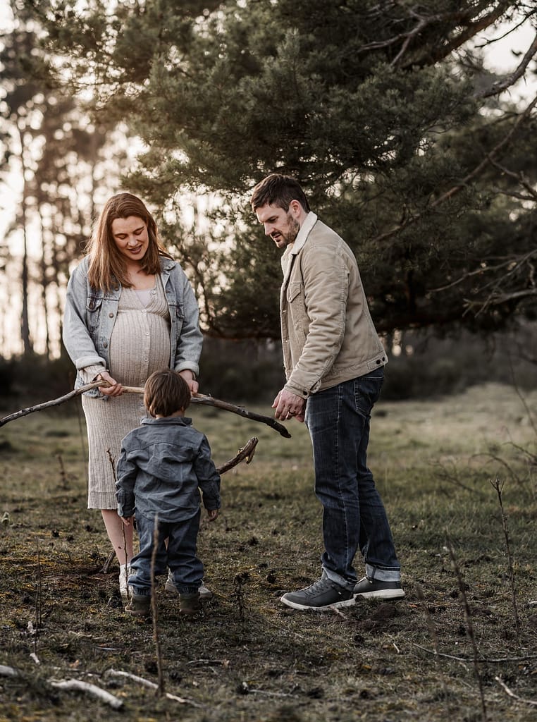 Familie mit kleinem Sohn und schwangerer Mama im Wald. Sie spielen zusammen. Der Sohn bringt Mama einen großen Ast. Im Hintergrund geht die Sonne golden unter.