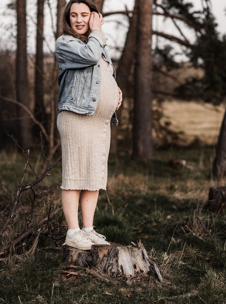 Schwangere Frau steht im Wald und schaut verträumt in die Ferne, während sie sich über den Babybauch streichelt.