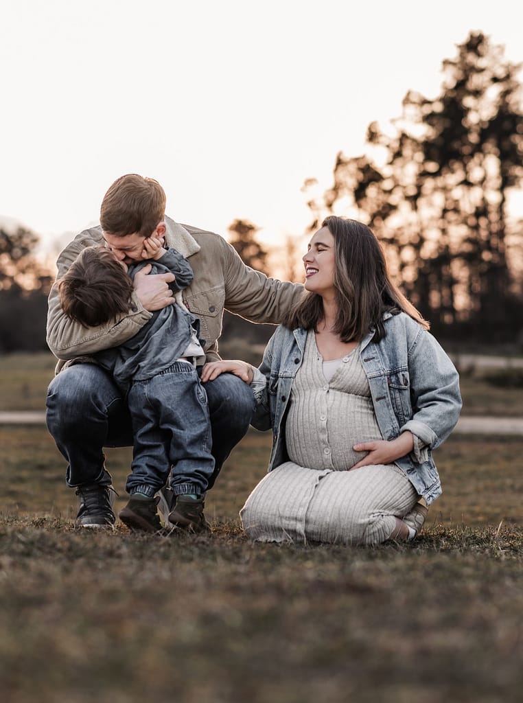 Familie mit kleinem Kind beim Babybauch Shooting in der Natur in Nuernberg und Fürth. Sie lachen gemeinsam und knien auf einer Wiese beim Sonnenuntergang.