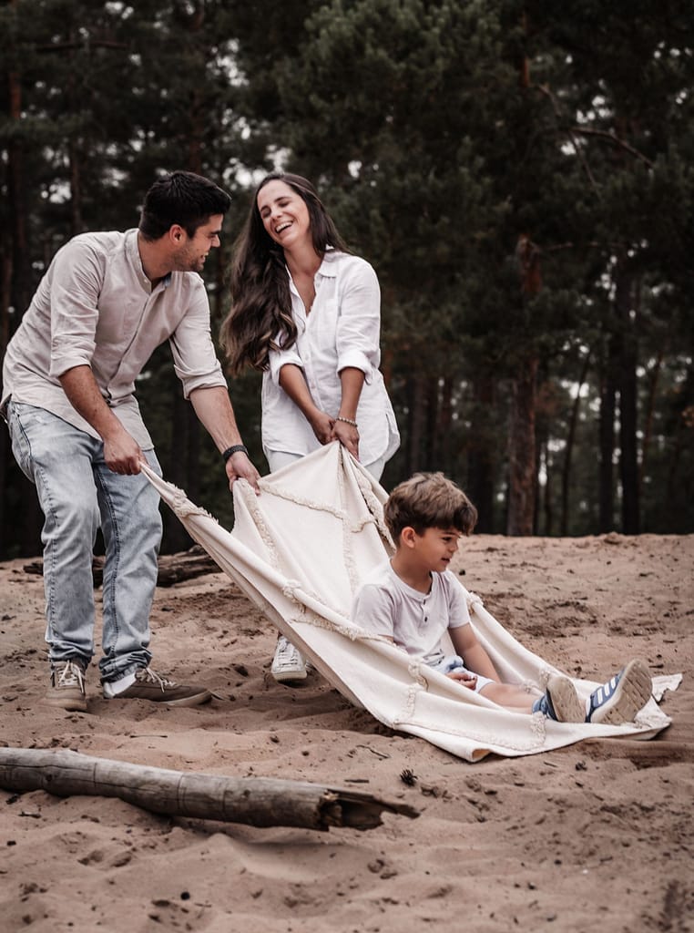 Familie mit zwei kleinen Kindern beim Familienshooting auf der Sanddüne ind Nürnberg. Sie ziehen ihren 5-jährigen Sohn mit einer Picknickdecke über den Sand. Ein kleines Mädchen krabbelt hinterher.